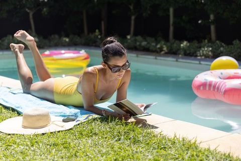 Woman sunbathing and reading paperback by backyard pool in yellow swimsuit