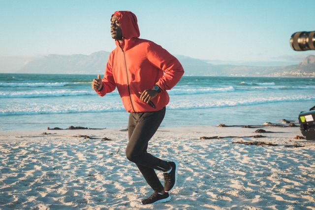 African American man running on a beach at sunset, wearing a red hoodie and black pants. The ocean waves and mountains in the background create a scenic view. Ideal for promoting fitness, healthy lifestyle, outdoor activities, and exercise routines.
