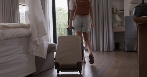 African american woman walking to balcony pulling suitcase in bright hotel bedroom