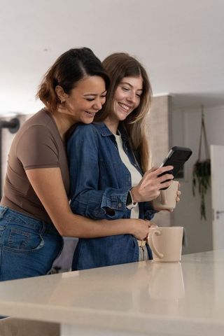 Two women embracing and checking smartphone while holding mugs at modern kitchen island