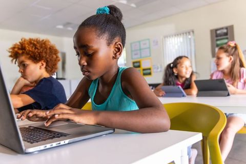 African American girl typing on laptop in classroom with diverse classmates using tablets