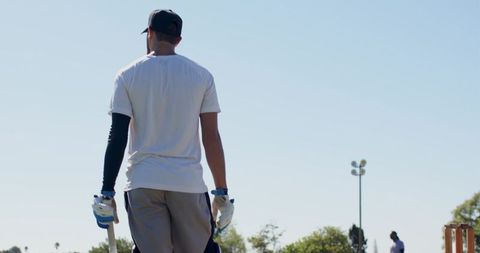 Male cricketer walking to pitch holding bat and gloves under bright sunny sky