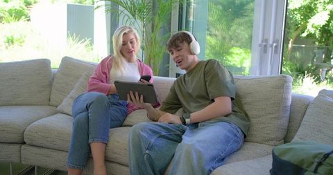 Mother and teen son relaxing on sofa with tablet and headphones in sunlit living room