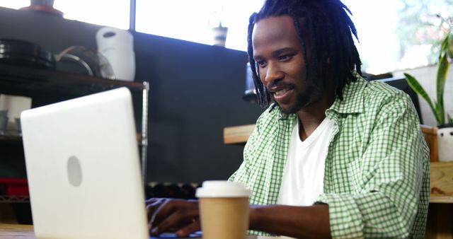 Close-up of smiling man with dreadlocks and plaid shirt working on laptop in a casual cafe environment. Coffee cup in foreground, while natural light from window illuminates the workspace. Perfect for depicting modern technology usage, remote work, freelancer lifestyle, coworking spaces, and casual professional settings.