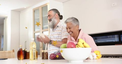Laughing mature couple cooking together in bright modern kitchen with fresh fruit bowl