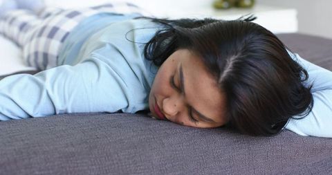 Asian woman lying on gray bed wearing blue top and plaid pajama pants, resting and napping