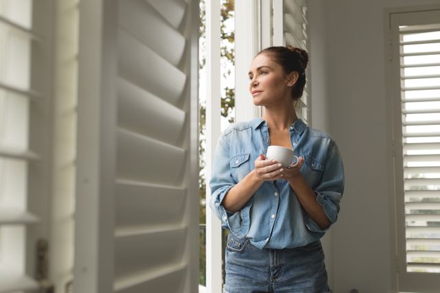 Woman standing by window holding coffee cup, dressed in casual denim shirt and jeans. Natural light streaming in, creating a peaceful and relaxed atmosphere. Ideal for use in lifestyle blogs, home decor websites, or advertisements promoting relaxation and comfort at home.