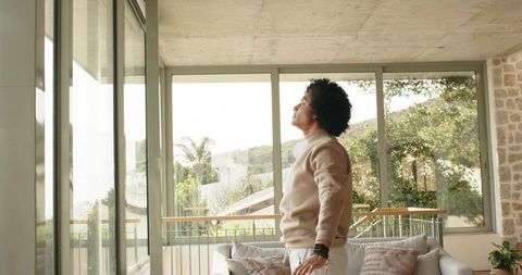 Man in thirties standing by sunlit balcony window wearing beige sweater, reflecting