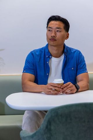 Asian man sitting at minimalist cafe bench holding takeaway coffee and looking thoughtful