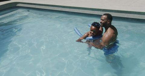 African american dad holding son in backyard pool with noodle and goggles, summer bonding