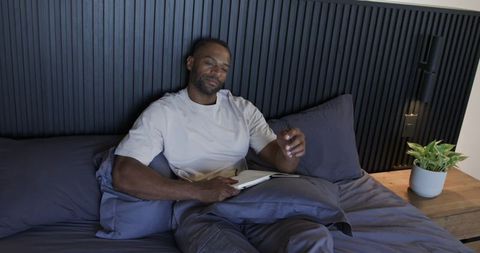 Man reclining in bed reading journal and holding glasses in modern bedroom with plant