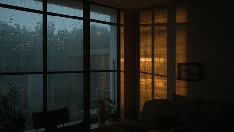 Rain-streaked floor-to-ceiling window casting warm golden light into moody living room