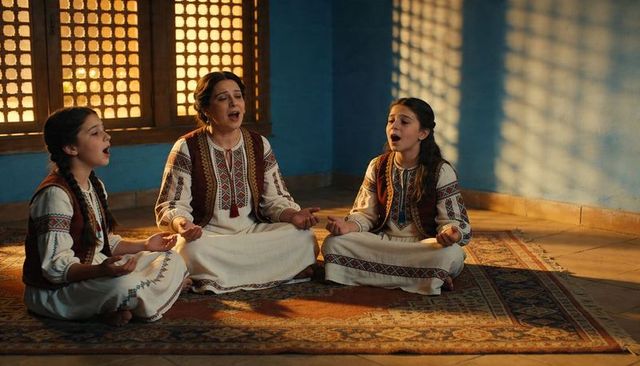 Sunlit mother and daughters singing in embroidered folk dresses and vests near mashrabiya window