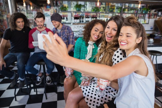 Group of cheerful friends enjoying time together in a retro-style diner. Three women in the foreground are taking a selfie while holding milkshakes, smiling and having fun. In the background, two men are sitting and chatting. Perfect for themes related to friendship, socializing, youth culture, and leisure activities.