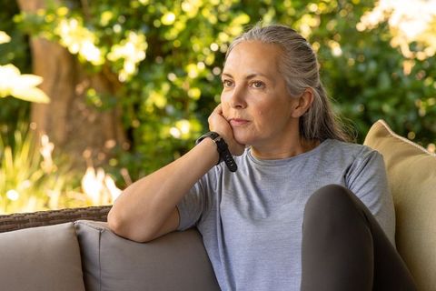 Mature woman sitting on patio rattan sofa resting head wearing fitness tracker gazing