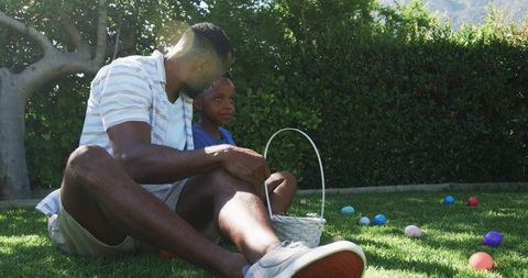 African american father and teenage son sitting on lawn during backyard egg hunt