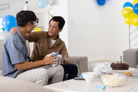 Asian father and teen son sharing wrapped birthday gift in cozy living room celebration