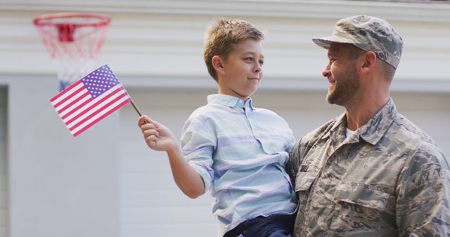 This image shows a military father smiling while holding his son, who is waving an American flag. They are standing outside near a house, and there's a basketball hoop in the background. This image is ideal for themes related to military families, patriotism, homecoming celebrations, or Father's Day promotions.