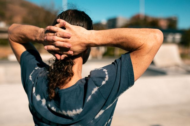 Back view of a man standing at a skatepark with his hands crossed on his head, enjoying a sunny day. Ideal for use in lifestyle blogs, urban recreation promotions, and summer activity advertisements.