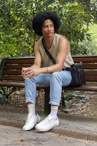 Young man sitting on park bench holding coffee cup gazing thoughtfully wearing crossbody bag
