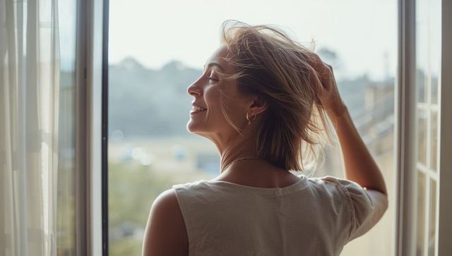 Woman enjoying morning breeze at open balcony door, backlit profile and relaxed smile