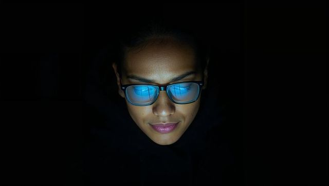 Woman in hood gazing down at blue screen reflection in glasses closeup moody low-light