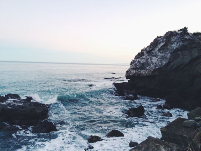 This image captures a tranquil and scenic view of a rocky coastline at dusk with calm ocean waves gently hitting the shoreline. The horizon is subtly illuminated by the fading light, creating a peaceful and picturesque setting ideal for promoting travel destinations, relaxation, and nature appreciation. Perfect for use in tourism brochures, inspirational content, and mindfulness visuals.