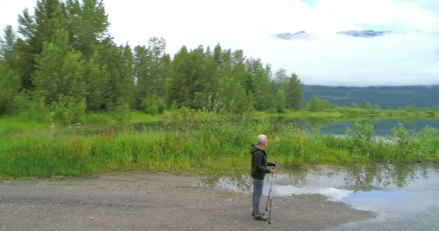 A senior man standing near a tranquil lake with hiking poles, enjoying the natural scenery. Trees and greenery surround the calm water, reflecting a peaceful atmosphere. This image is ideal for promoting outdoor activities, relaxation in nature, and adventures for elderly individuals. It can be used in travel brochures, fitness campaigns, health and wellness websites, and senior lifestyle magazines.
