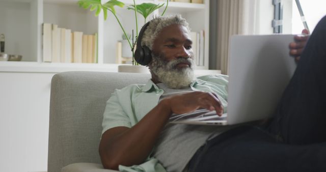 Mature man with gray beard relaxing on sofa while using laptop and wearing headphones. He appears comfortable in casual attire with bookshelves and indoor plant in background, suggesting a home office or casual work environment. This can be used for promoting remote work lifestyles, relaxation, technology, home environments, and leisure activities.