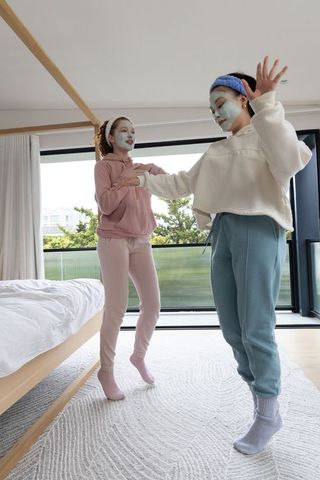 Teen friends wearing facial masks dancing and holding hands in bright modern bedroom