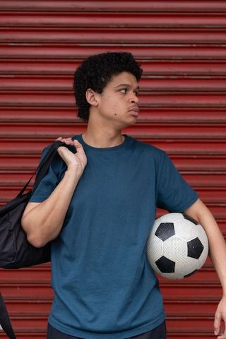 Young african american man holding soccer ball carrying duffel against red shutter