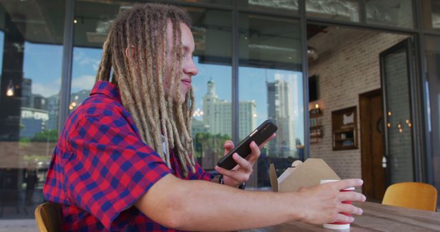 Young man in a red checkered shirt using his smartphone while holding a cup of coffee at an outdoor cafe table. This image is perfect for promoting urban lifestyle, relaxation, technology use, or cafe culture. Ideal for advertisements, websites, or blogs related to coffee shops, mobile apps, or leisure activities.