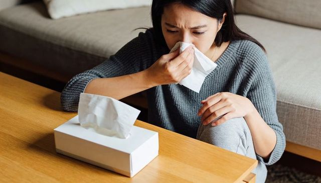 Young asian woman sneezing into tissue at home living room holding tissue feeling unwell