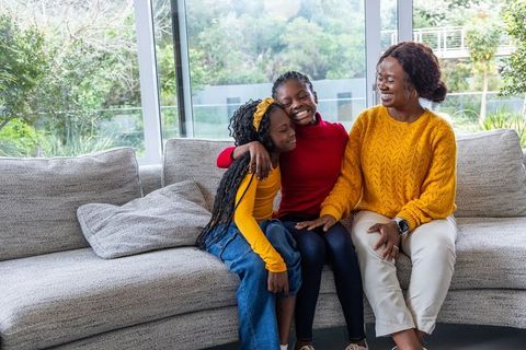 African american mother and daughters sharing joyful hug on sunlit modern living room sofa