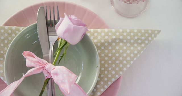 A neatly presented table setting features a pastel-themed plate and napkin arrangement, adorned with a delicate pink rose and cutlery tied with a ribbon, with copy space. Such a setup suggests a special occasion or a celebration, emphasizing attention to detail and a touch of elegance.