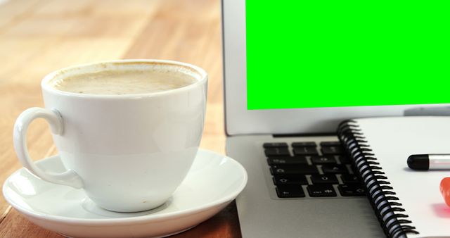 Cup of coffee sitting beside laptop with green screen and notebook laying on desk. Perfect for illustrating home office setups, remote working environments, productivity, and modern workspaces.