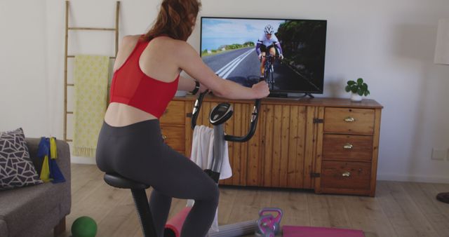 A woman exercising on a stationary bike in a living room while watching a virtual cycling tour on the TV. She wears a red sports bra and gray leggings. Perfect for illustrating home fitness, indoor workouts, and maintaining an active lifestyle at home.