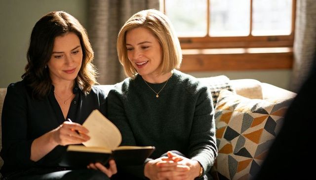 Two women sharing notebook on sofa, reading and smiling together in cozy living room