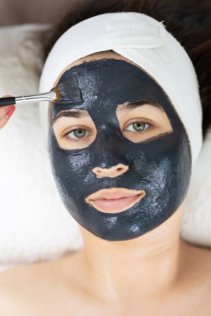 Beautician applying black face mask to female customer in a beauty salon. Customer lying down with a headband holding hair back, receiving a facial treatment aimed at skincare and relaxation. Ideal for use in beauty, skincare, and wellness industry promotions, advertisements for beauty salons, and content about self-care and luxury beauty treatments.