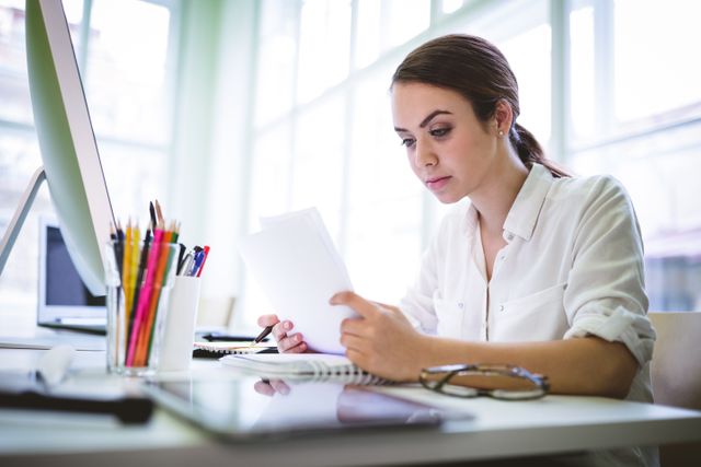 Young female graphic designer concentrating on reviewing documents at her office desk. Ideal for use in articles or advertisements related to creative professions, office environments, productivity, and professional work settings.