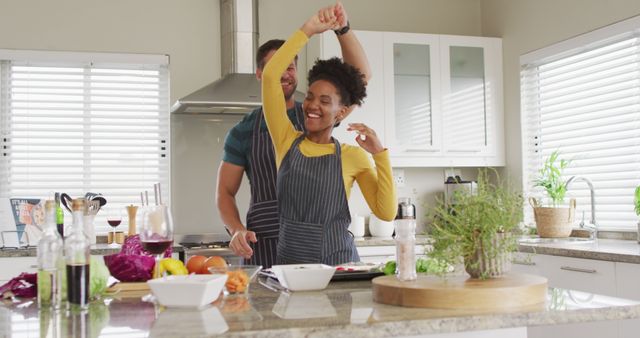 Happy couple dancing and cooking together in a modern kitchen. They both wear aprons while joyfully preparing food. Great for promoting related products or services such as home cooking, lifestyle, family values, relationships, or culinary tools.