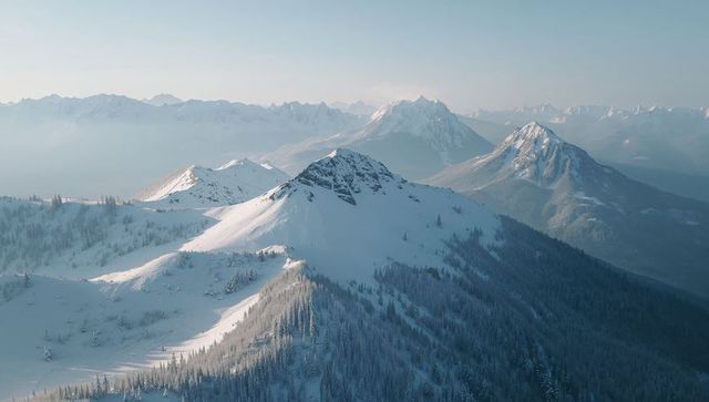 Sunlit alpine ridge rising through morning haze with snow-capped peaks and conifer forest