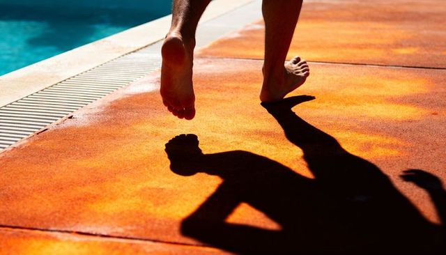 Stepping barefoot along orange pool deck casting long shadow over blue water