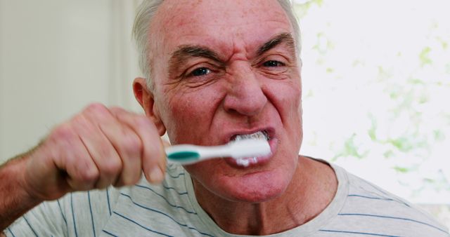 Elderly man brushing teeth with a serious expression, demonstrating the importance of dental hygiene for seniors. Can be used for health care promotions, dental products, articles on senior health, and oral care education.