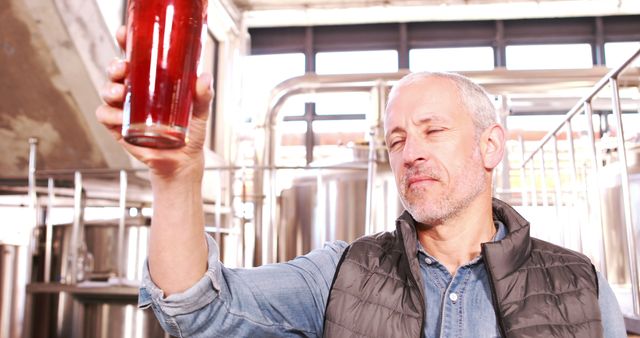 Senior man, an experienced brewer, is inspecting a glass of craft beer in a modern brewery. Suitable for use in articles or promotional content related to brewing techniques, craft breweries, beverage quality control, and the beer industry.