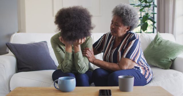 Grandmother sitting on a couch comforting her sad granddaughter who has her head in her hands. Cups on coffee table suggest a casual home environment. Ideal for use in articles or presentations on family support, emotional wellness, intergenerational relationships, and mental health.