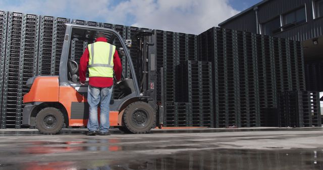 Worker in high-visibility safety vest operating forklift in an industrial warehouse area with numerous stacked pallets. Suitable for articles, advertisements, and content related to logistics, warehouse safety, manual labor in industrial settings, and operational management.