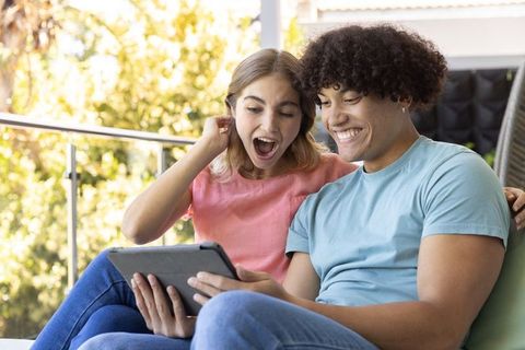 Young couple laughing with tablet on sunlit balcony, relaxing outdoor romantic moment