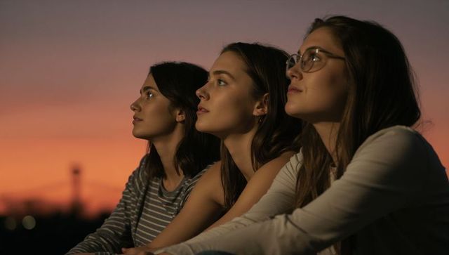 Three young women gazing at sunset on rooftop railing during golden twilight skyline