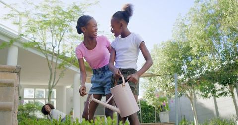 Smiling teenage girls watering garden and tending raised bed on sunny backyard patio